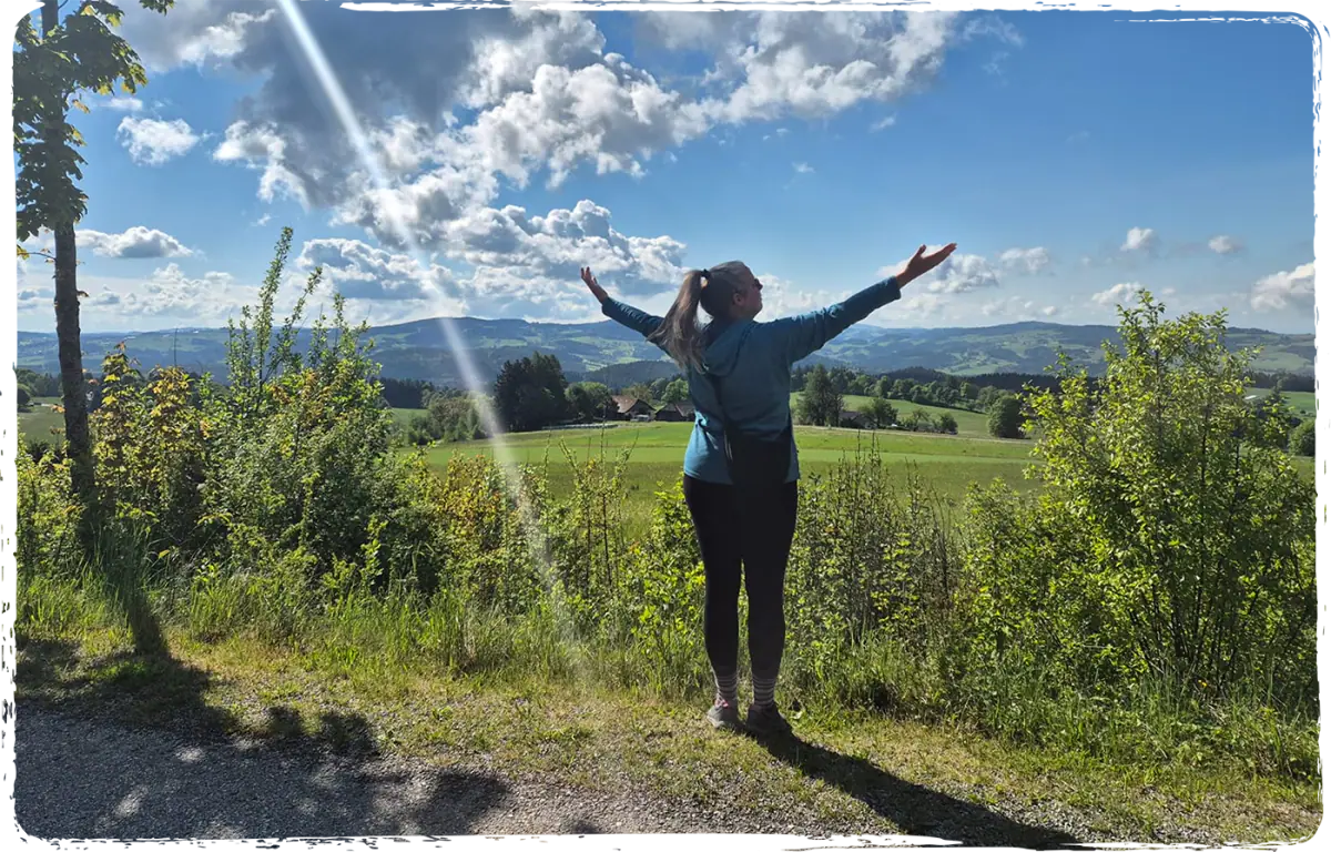 Raphaela Jóia Jordão steht mit ausgebreiteten Armen vor einer grünen Hügellandschaft